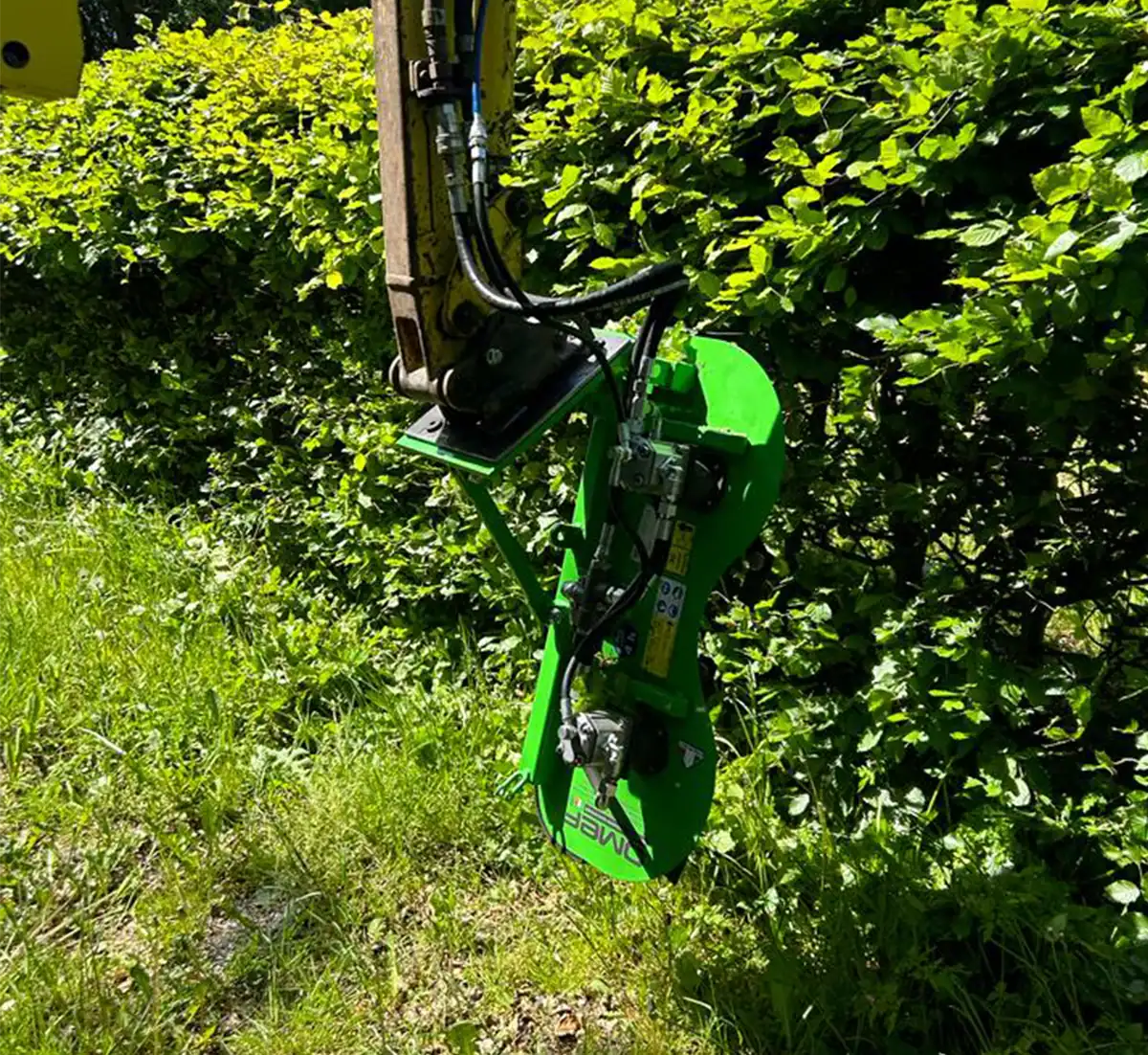 OMEF TG series hydraulic hedge trimmer on an excavator arm, its disc head set vertically against a tall beech hedge