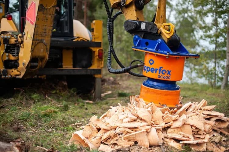 Orange and blue Dipperfox SC600 stump grinder on a backhoe boom, standing over the pile of coarse wood chips it has just produced