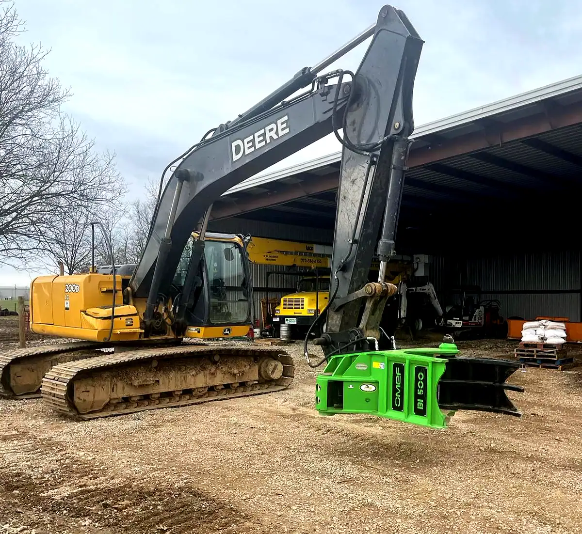 OMEF BIG INCH BI500 tree shear in green, mounted on the stick of a John Deere 200D LC excavator in a contractor's yard